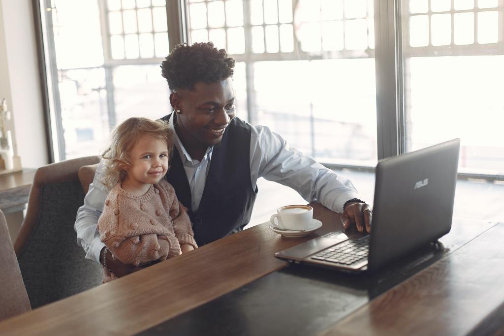 Homem trabalhando em seu notebook com sua filha no colo, em uma ação de sua empresa para o dia dos pais.