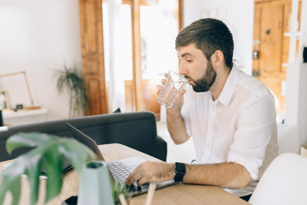 Homem sentado em uma mesa lendo em seu notebook sobre a regulamentação do home office enquanto toma um copo de água.